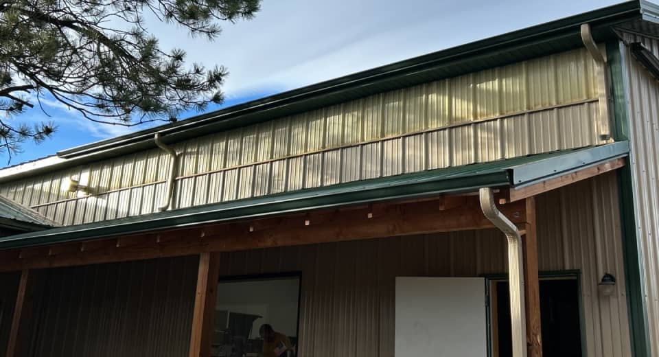 Exterior of a metal building with green trim, wooden beams, and a large window. A tree branch extends into the frame under a partly cloudy sky.